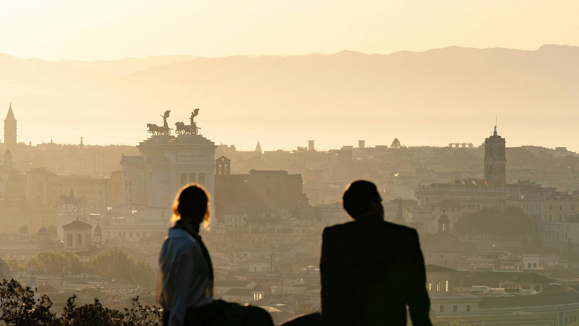 Two silhouetted figures gazing over a hazy golden Rome skyline at dawn