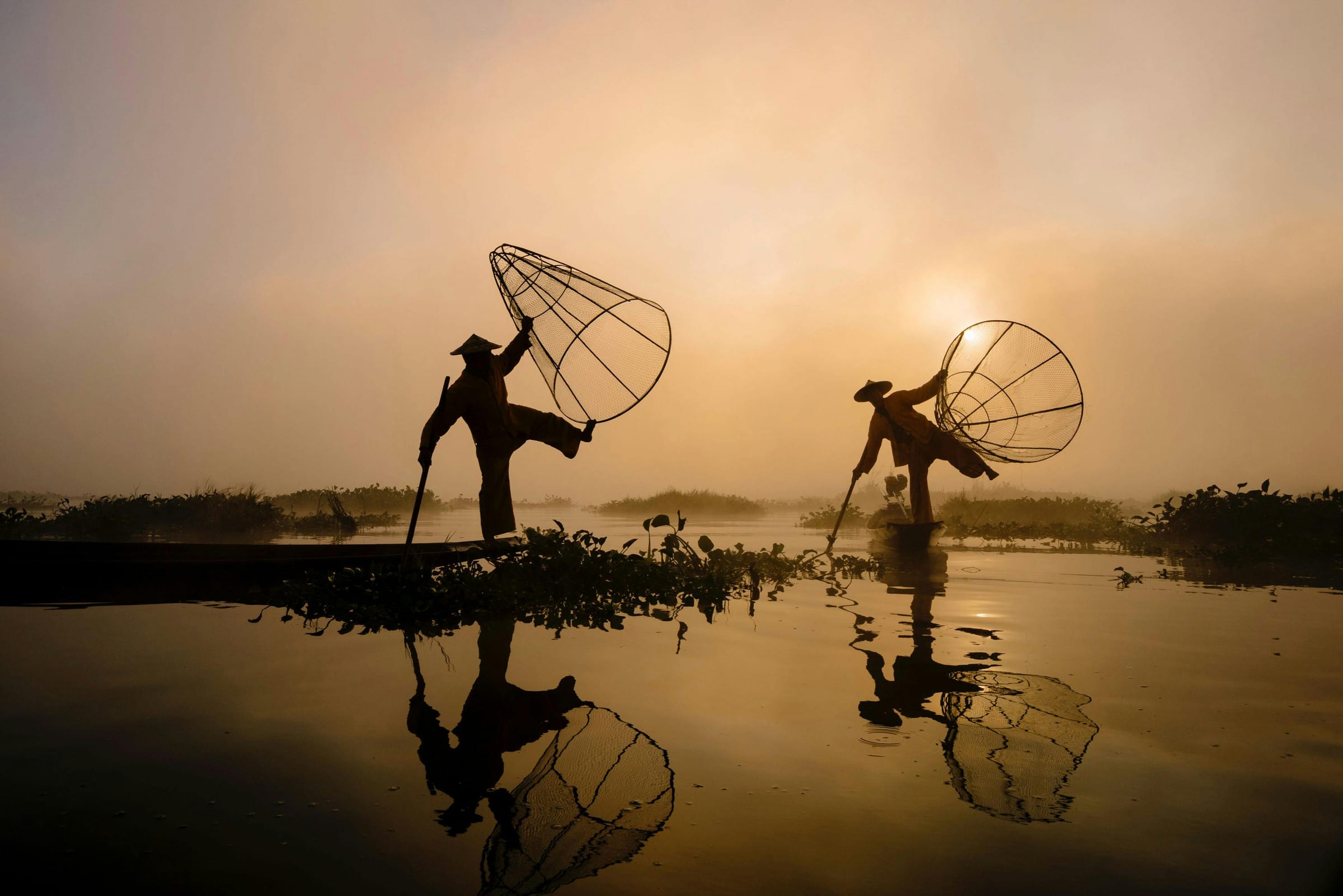 Two fishermen with conical nets silhouetted on a misty lake at golden dawn