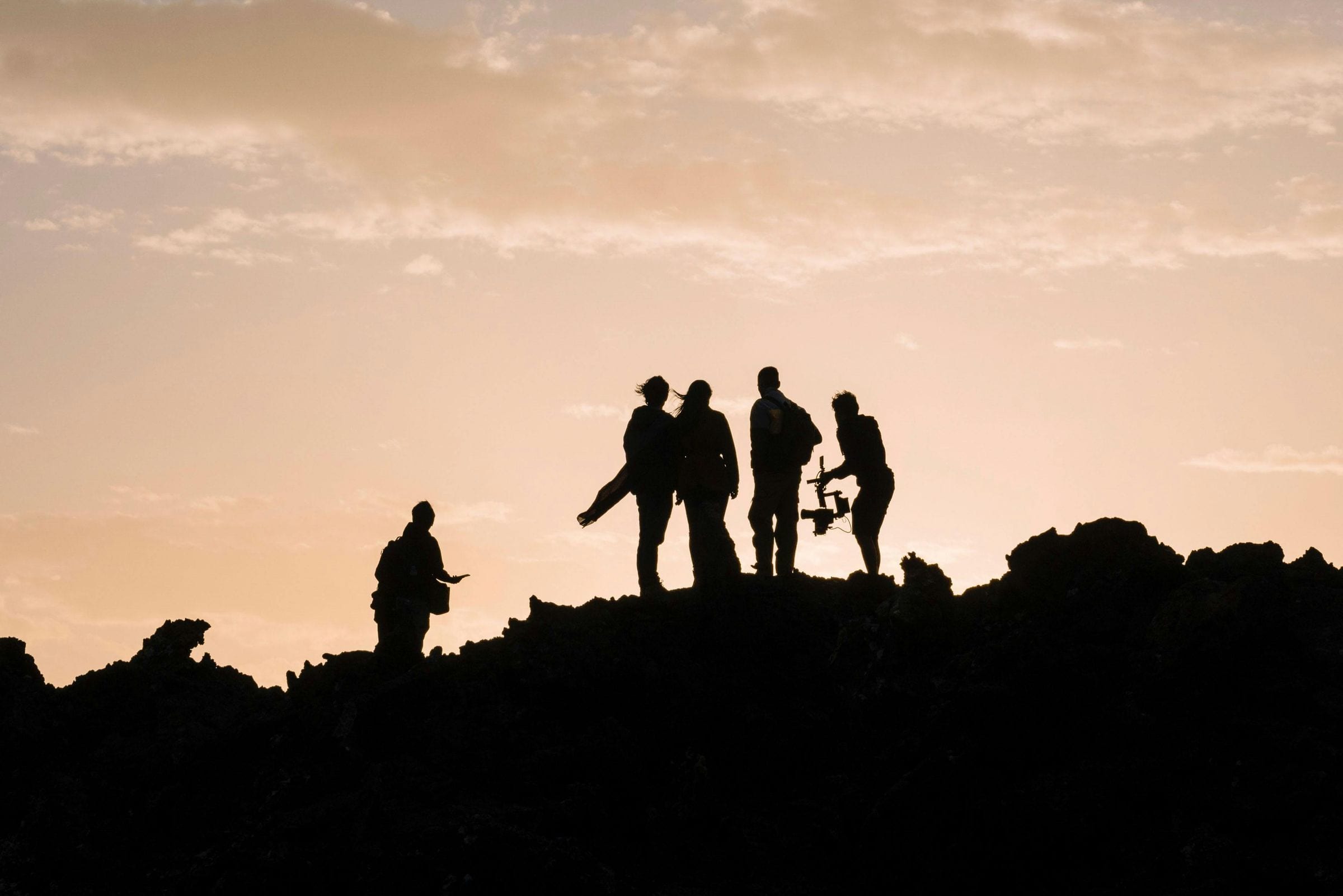Film crew silhouetted on a rocky ridge at sunset, one operating a gimbal rig