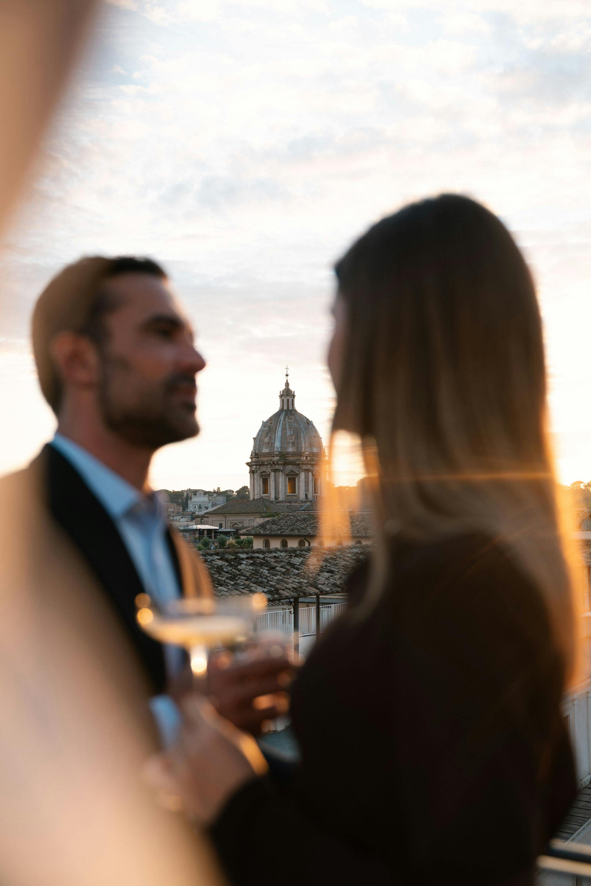 Couple with drinks on a rooftop terrace at dusk, a Roman church dome beyond