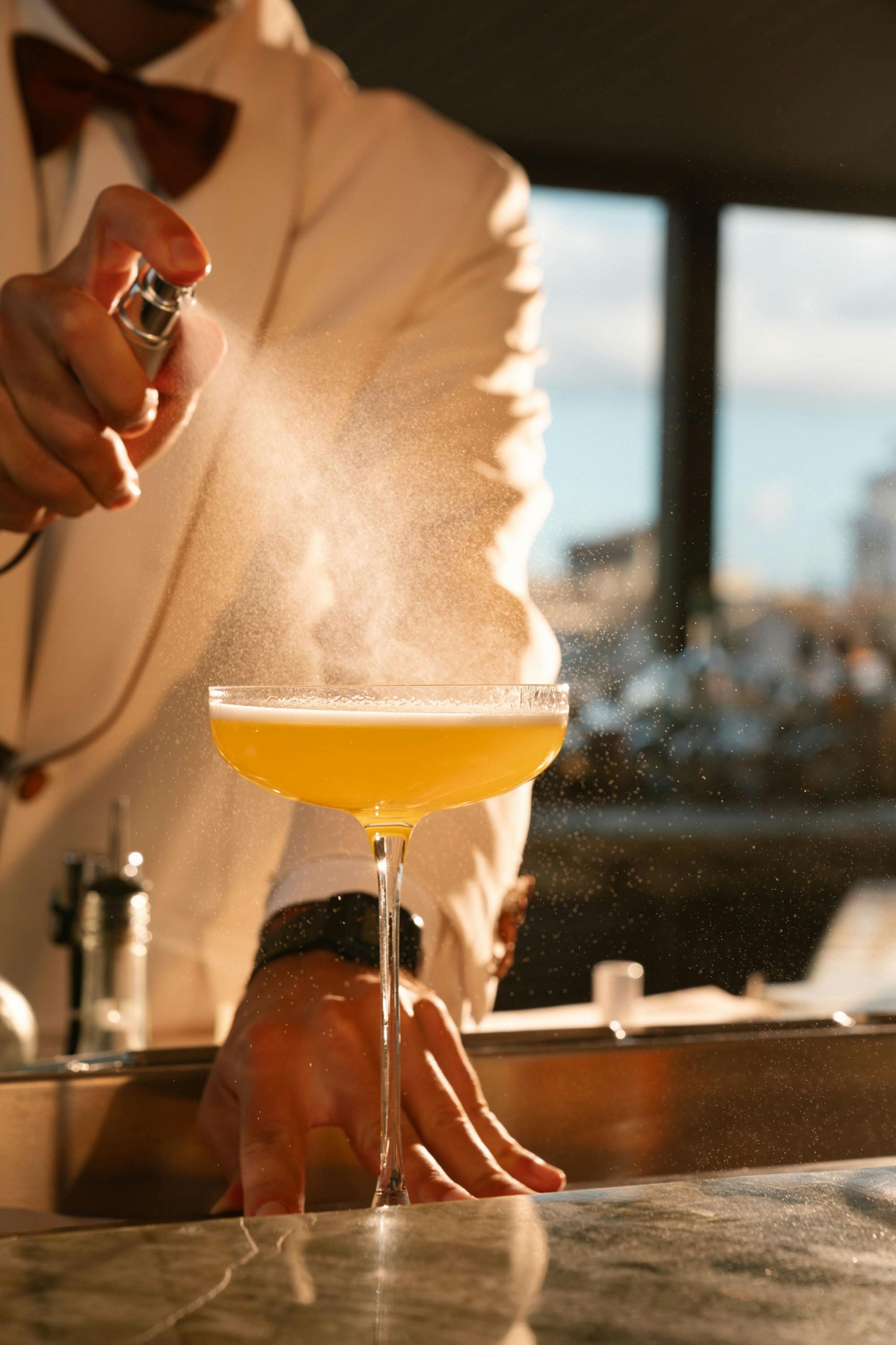 Bartender misting a citrus cocktail in a coupe at a sunlit rooftop bar