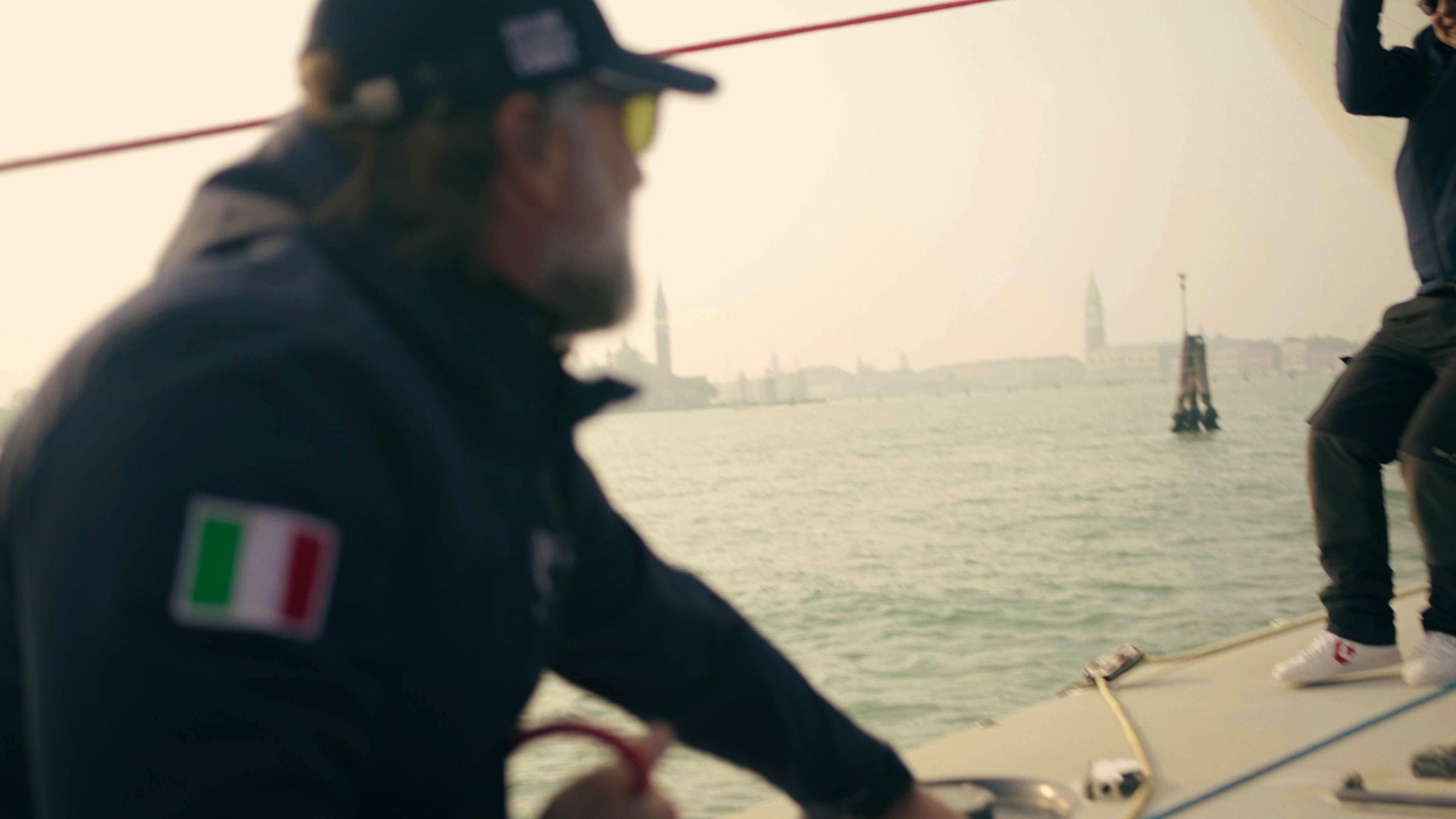 Crew member with Italian flag patch at the helm, hazy Venice skyline behind